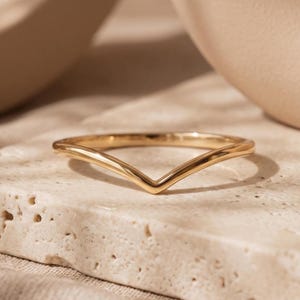 May include: A polished gold ring featuring a V-shaped chevron design. The band has a smooth, reflective surface and a pointed centre, creating a distinct angular silhouette. The metal has a warm, yellow-gold tone with a high-shine finish.