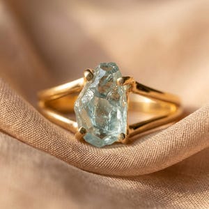 Puede incluir: Un anillo de oro con una piedra preciosa de aguamarina en bruto de color azul claro sujeta por cuatro garras. La banda del anillo es lisa y pulida, con un diseño dividido que se conecta al engaste que sostiene la piedra de cristal irregular.