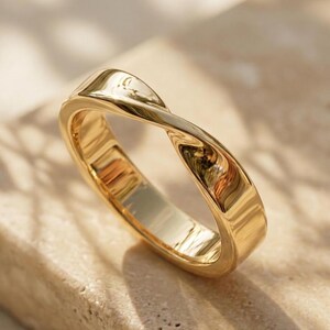 Puede incluir: Un anillo de oro pulido con un diseño trenzado continuo que forma una figura similar al infinito. La banda tiene una superficie lisa y reflectante con un perfil redondeado, mostrando una construcción de bucle sin costuras.