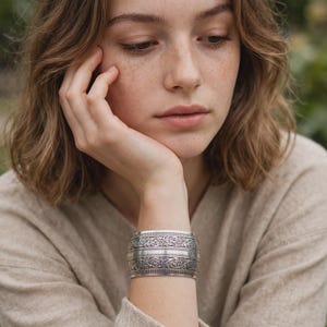 May include: A wide silver cuff bracelet featuring intricate embossed floral scrollwork patterns across the surface, with a central horizontal band of fine textured detailing that runs along the entire circumference of the metal wrist accessory.