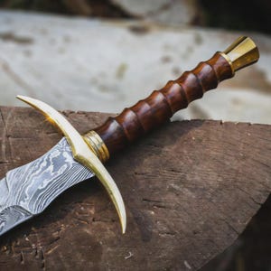 May include: A dagger featuring a patterned Damascus steel blade with a wavy, layered appearance. The hilt is crafted from dark, polished wood with a ribbed, cylindrical grip, accented by a bright gold-coloured metal crossguard and pommel.