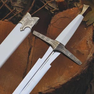 May include: A sword with a white blade and handle, featuring a metallic crossguard with intricate engraved patterns. The sword is accompanied by a matching white scabbard that has a metallic throat piece decorated with detailed scrollwork.