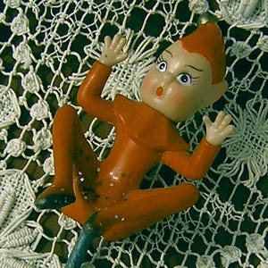 May include: A vintage orange pixie figurine with a surprised expression, mounted on a green metal stick. The pixie is lying on a white lace doily with floral patterns.