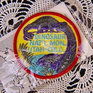 May include: A round patch with a dinosaur design and the text "DINOSAUR NAT'L MON. UTAH-COLO." is displayed on a yellow background with a red border. The patch is on a white doily.
