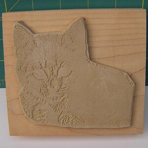 May include: A light brown cat-shaped rubber stamp on a rectangular wooden block. The stamp shows a detailed relief of a cat's face and body, with visible fur texture. The wooden block has a natural wood grain.