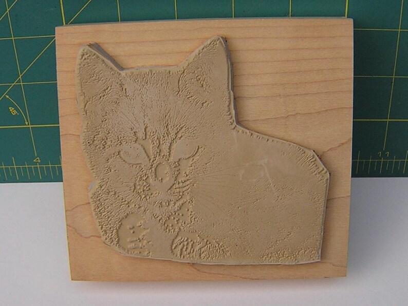 May include: A light brown cat-shaped rubber stamp on a rectangular wooden block. The stamp shows a detailed relief of a cat's face and body, with visible fur texture. The wooden block has a natural wood grain.