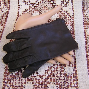 May include: A pair of dark brown leather gloves with a smooth finish and stitched detailing along the fingers, displayed on a pale mannequin hand form to show the shape and structure of the wrist-length design.