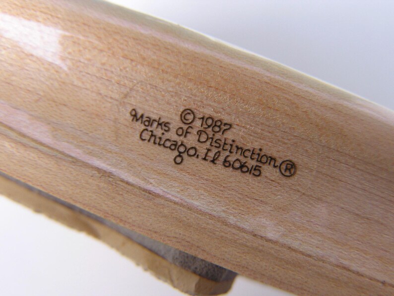 May include: Close-up of a wooden object with visible grain. The text "Marks of Distinction &copy; 1987 Chicago, IL 60615" is etched into the wood. The object appears to be a part of a larger wooden item.