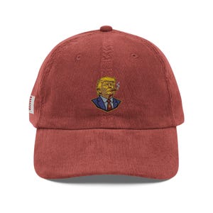 Peut inclure: Une casquette de baseball en velours côtelé rouge avec une broderie représentant une personne aux cheveux blonds portant un costume bleu et une cravate rouge, fumant un cigare. Un petit drapeau américain est visible sur le côté.
