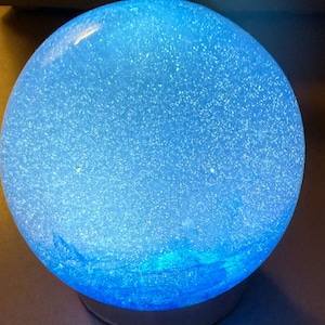 May include: A spherical lamp emitting a bright blue light, featuring a textured surface filled with numerous tiny, reflective glitter particles that create a shimmering effect across the entire round body of the illuminated object.