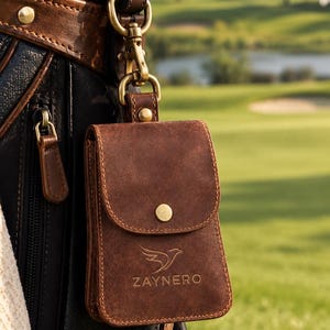 Könnte beinhalten: Eine braune Ledertasche mit Klappverschluss und einem Messing-Druckknopf, die den geprägten Text ZAYNERO und ein Vogel-Logo aufweist. Die Tasche ist mit einem Messing-Drehclip und einem passenden Lederriemen an einer schwarzen Golftasche befestigt.