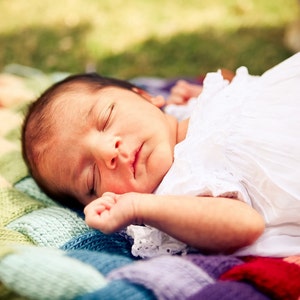 May include: A sleeping baby wearing a white dress lies on a colorful, rainbow-patterned knitted blanket. The blanket features squares of red, orange, yellow, green, blue, and purple. The baby has dark hair and is resting peacefully.