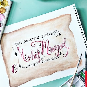 May include: Watercolor painting on paper with the phrase "I Solemnly Swear I'm Up To No Good" in burgundy script. The paper has a parchment-like appearance. Paintbrushes, watercolor paints, and an eraser are visible.
