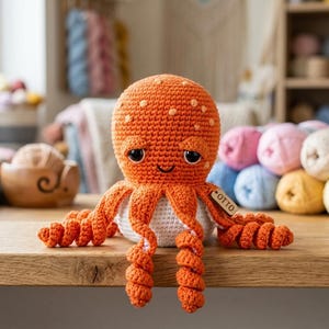 Peut inclure: Une peluche pieuvre crochetée avec une tête orange vif ornée de taches orange clair, de grands yeux noirs et un petit sourire brodé. Le dessous est blanc et elle possède huit tentacules orange bouclés avec une étiquette en bois marquée OTTO.