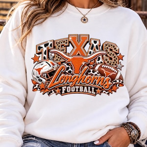 Peut inclure: Un sweat-shirt blanc à manches longues avec un motif graphique portant le texte TEXAS LONGHORNS FOOTBALL. Le design comprend une silhouette de bœuf longhorn, un casque de football américain et un ballon, avec des accents orange, noir et léopard.