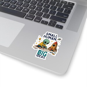 May include: A die-cut sticker featuring a green alien wearing a yellow hoodie, drawing on paper next to a small rocket ship. The design includes stars, a lightbulb, and a planet, with the text SMALL HUMAN. BIG IDEAS. in dark blue and teal.