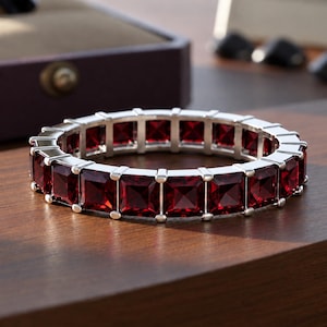Könnte beinhalten: Ein kreisförmiges Armband mit einer durchgehenden Reihe quadratisch geschliffener, tiefroter Edelsteine in einer Fassung aus poliertem, silberfarbenem Metall. Das Design besteht aus mehreren identischen quadratischen Gliedern.