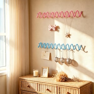 Könnte beinhalten: Zwei horizontale Wandhalterungen mit einem zickzackförmigen, geschwungenen Drahtdesign. Die obere Halterung ist rosa und hält eine rosa Haarschleife sowie eine silberne Stern-Halskette. Die untere Halterung ist blau und hält eine Karte und drei Teesiebe.