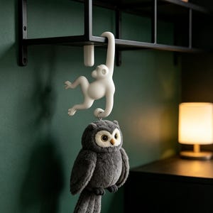 May include: A white monkey-shaped hook hanging from a black metal shelf, with a plush dark gray owl toy suspended from the monkey's tail. The owl features large light-colored eyes, a small beak, and soft fur, hanging vertically from the hook.