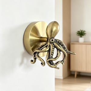 May include: A brass-toned metal wall hook shaped like an octopus. The hook features a circular mounting plate and a detailed octopus figure with curved tentacles extending outward, showcasing a textured finish with visible suction cup details.