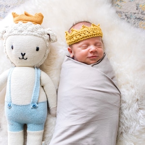 May include: A newborn baby wearing a yellow knitted crown sleeps peacefully in a gray swaddle. A white knitted lamb with blue overalls sits beside the baby.