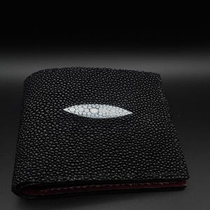 May include: A black bifold wallet featuring a textured surface with a prominent, smooth, white, almond-shaped inlay centered on the front panel. The wallet has a dark, deep red interior lining visible along the bottom edge of the folded structure.