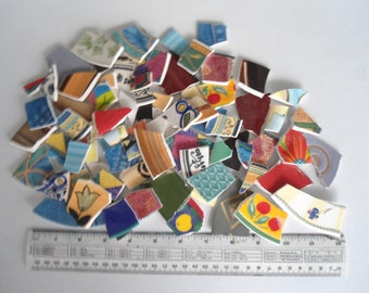 May include: A pile of colorful, broken ceramic tiles with a ruler for scale. The tiles are various shapes and sizes and feature floral, geometric, and abstract designs.