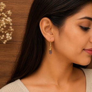 May include: A gold drop earring featuring a small, textured gold cap at the top and a dangling, elongated, translucent purple gemstone pendant with a faceted surface. The earring hangs from a thin gold wire hook attached to the earlobe.