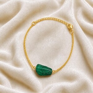 May include: A gold-toned chain bracelet featuring a single, raw, irregularly shaped green stone as a central focal point. The bracelet includes a delicate link chain and a small lobster claw clasp closure, showcasing a minimalist design.