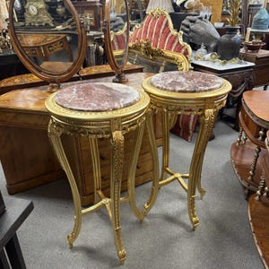May include: A pair of matching round side tables featuring circular tops with reddish-brown marble inlays. The tables have ornate gold-finished frames with curved legs, intricate carved detailing along the edges, and a cross-stretcher base.