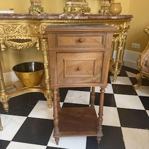 May include: A wooden bedside table featuring a dark marble top, one small upper drawer, and one larger lower cabinet door. The piece stands on four turned legs connected by a lower shelf, with each drawer and the cabinet door featuring a small knob.