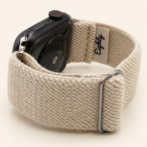 May include: A black smartwatch with a textured, off-white woven fabric band. The band features a small rectangular tag with the word "Eighty" printed in black script and a silver-toned metal buckle for adjustment and secure fastening.