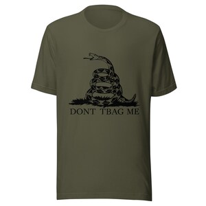 Könnte beinhalten: Ein olivgrünes Kurzarm-T-Shirt mit einer schwarzen Grafik einer zusammengerollten Klapperschlange auf einem Grasbüschel. Unter der Schlange ist der Text DONT TBAG ME in einer schwarzen Serifenschrift gedruckt. Das Shirt hat einen Rundhalsausschnitt.