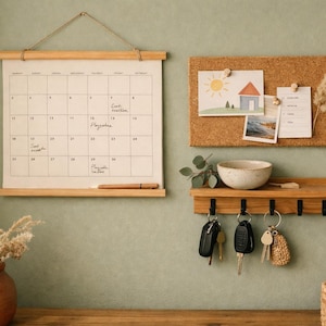 Op de afbeelding: Een wandkalender van stof met houten rails, een rechthoekig prikbord van kurk met opgeprikte notities en tekeningen, en een houten plank met vijf zwarte metalen haken waaraan sleutels en een kleine keramische kom hangen.
