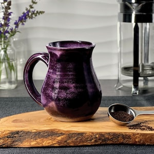 May include: A deep purple ceramic mug with a glossy, mottled finish and a curved handle. The mug has a rounded body that tapers toward the rim, featuring a lighter, natural-toned base. The surface displays variations in color intensity.