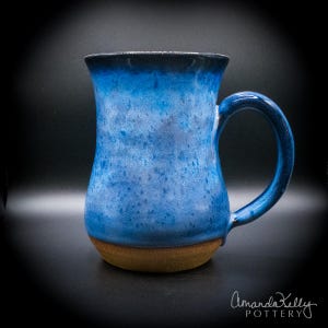 Puede incluir: Una taza de cerámica con un esmalte azul vibrante y texturas moteadas que transicionan hacia un borde azul marino más oscuro. La taza tiene un asa curva y una abertura ancha, con una base de color marrón tierra sin esmaltar. El texto Amanda Pottery es visible.