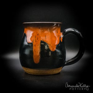 May include: A ceramic mug with a rounded body and a curved handle. The mug features a glossy black base glaze with a vibrant orange glaze dripping from the rim down the sides. The text Amanda Kelly Pottery is printed in white at the bottom right.
