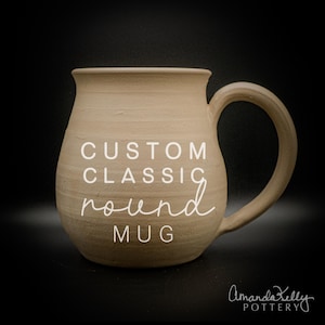 May include: A round, beige ceramic mug with a smooth, matte finish and a curved handle. The text CUSTOM CLASSIC round MUG is printed in white on the front, and the text Amanda Kelly POTTERY is printed in white in the bottom right corner.