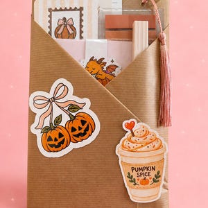 Può includere: Una busta di carta marrone contenente articoli di cancelleria, tra cui una lista di lettura, un adesivo a forma di zucca e un segnalibro a forma di drago. La busta è decorata con adesivi raffiguranti zucche e un latte speziato.
