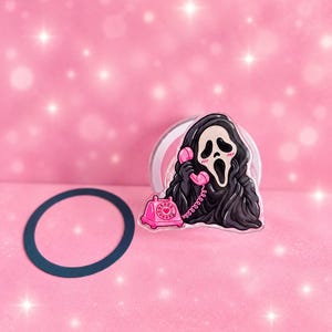 Op de afbeelding: Een acrylspeld met een in zwart gehulde figuur met een wit masker die een felroze draaitelefoon vasthoudt. De figuur heeft een wijd open mond en roze blosjes op de wangen. Een aparte donkere cirkelvormige ring ligt ernaast.