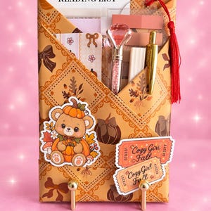 Può includere: Un organizer tascabile in carta color cuoio con motivo a fiocchi e zucche marroni, contenente una lista di lettura, una penna con punta a diamante, una penna dorata e foglietti adesivi. Presenta un adesivo con orsetto e due biglietti Cozy Girl Fall.