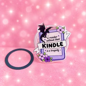 May include: A clear acrylic pin featuring a purple e-reader illustration with a black dragon, pink and purple flowers, and a white banner with the text: a reader without their KINDLE is a tragedy. A black circular ring is positioned nearby.