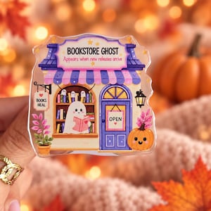 Op de afbeelding: Een doorzichtige acryl pin met een illustratie van een boekhandel met een geest die een boek leest. Het bord zegt BOOKSTORE GHOST Appears when new releases arrive, met een kleiner bordje BOOKS HEAL en een deurteken met OPEN. Een pompoen is zichtbaar.