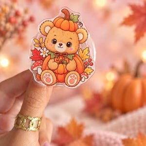 May include: An acrylic pin featuring a light brown cartoon bear sitting inside a large orange pumpkin. The bear wears a smaller pumpkin on its head and is surrounded by colorful autumn leaves, small white flowers, and red mushrooms.