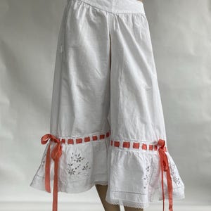 May include: White cotton culottes featuring a wide waistband, a vertical front slit, and a ruffled hem. The hem is decorated with eyelet embroidery, a scalloped lace edge, and a woven coral ribbon threaded through the fabric, tied into bows.