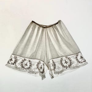 May include: Light gray shorts with a brown elastic waistband and a decorative border. The border features a repeating pattern of leaves and insects in a darker shade of gray. The shorts are made of a lightweight, semi-sheer fabric.