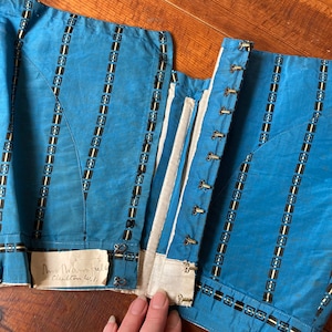 May include: A blue and white striped corset with a white cotton lining and metal eyelets. The corset has a handwritten note that says "Mrs. Hawthorne's custom 41.1/2".