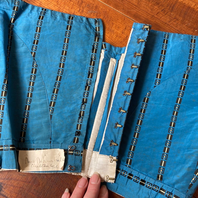 May include: A blue and white striped corset with a white cotton lining and metal eyelets. The corset has a handwritten note that says "Mrs. Hawthorne's custom 41.1/2".