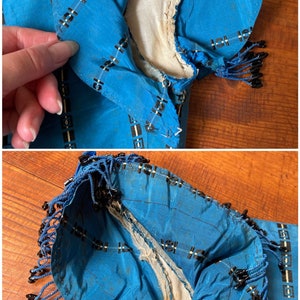 May include: A blue silk fabric with a black and white geometric pattern. The fabric has a fringe trim with black beads. The fabric is worn and has some tears.