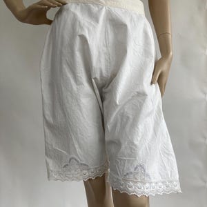 Könnte beinhalten: Weiße Baumwoll-Bloomers mit einem hoch taillierten Bund in einem helleren Cremeton. Die Beinöffnungen sind mit filigraner weißer Spitzenborte und zarten gestickten Blumendetails versehen, was für einen klassischen Look sorgt.